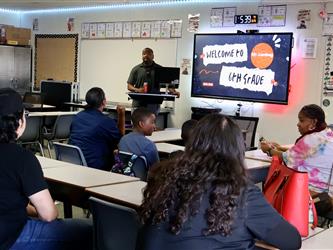 A teacher stands at the front of a classroom, welcoming 6th grade students and their parents.